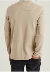 Beige Langarmshirt mit Rundhalsausschnitt, strukturiertem Stoff, Rückeneinsatz und flachen Nähten, getragen mit dunklen Jeans.
