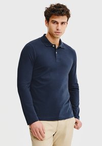 Granatowa koszula polo z długim rękawem, wykonana z teksturowanej tkaniny, z klasycznym kołnierzykiem i listwą z trzema guzikami. W zestawieniu z beżowymi spodniami.