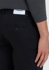 Zwarte broek van een duurzame stof, met een achterzak met een knoopdetail en een zichtbaar merklabel op de tailleband.