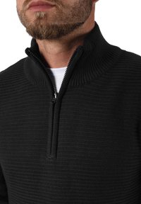 Schwarzer Zip-Hoodie aus strukturiertem, geripptem Stoff. Hoher Stehkragen und ein kurzer Reißverschluss betonen das Design.