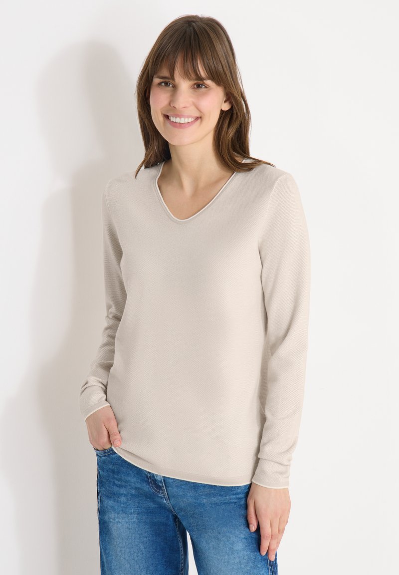 Femme aux cheveux bruns souriante, portant un haut à manches longues beige clair et un jean bleu, debout devant un fond blanc uni.