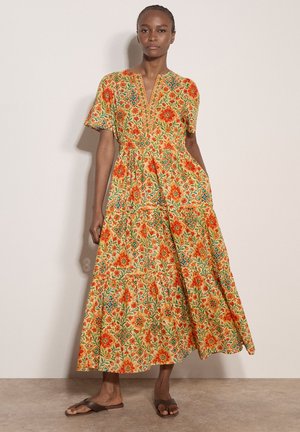 Frau trägt ein langes, kurzärmeliges Kleid mit orangefarbenen Blumen- und geometrischen Mustern, steht an einer schlichten hellen Wand und trägt braune Sandalen.