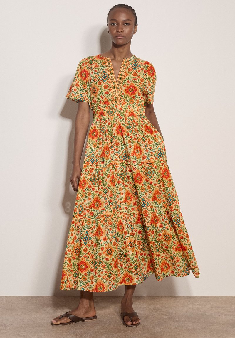 Woman wearing a long, short-sleeve dress with orange floral and geometric patterns, standing against a plain light wall, wearing brown sandals.