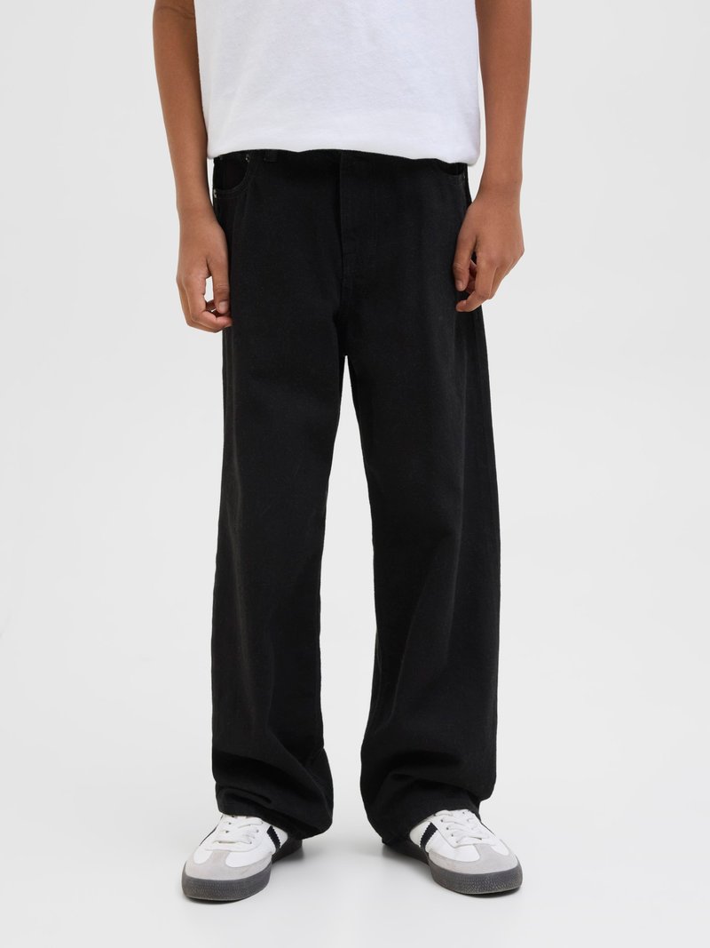 Person wearing loose black pants, white t-shirt, and white sneakers with gray soles, standing against a plain light background.