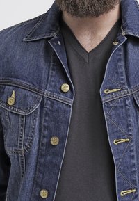 Tummansininen denim-takki, jossa on kaksi rintataskua, hopeiset napit ja keltaiset ompeluyksityiskohdat; käytetään mustan V-pääkappaleen päällä.
