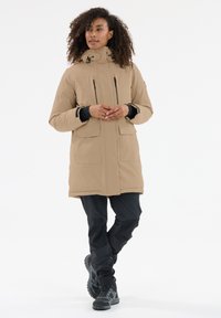 Beige vaatteiden eristysohjaus, jossa on huppu, vetoketjullinen etuosa ja kaksi etutaskua. Asusteena mustat housut ja tukevat mustat saappaat.