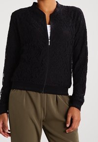 Bomber jacket in pizzo nero con zip frontale, polsini e orlo a costine. Motivo floreale intricato; tessuto leggero e strutturato.