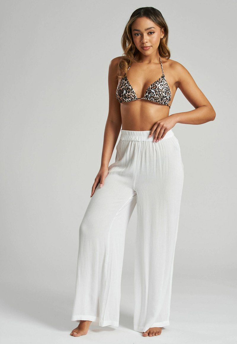 Top de bikini triangular de estampado de leopardo combinado con pantalones amplios, blancos y texturizados. La modelo posa con una mano en la cadera y descalza.