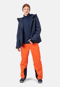 Navy wasserfeste Jacke über einem navy-farbenen Fleece, kombiniert mit leuchtend orangefarbenen Schneehosen mit schwarzen Akzenten. Das Model trägt navy-farbene Schuhe und eine gemusterte Mütze.