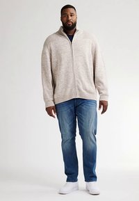 Svetle béžový svetr na zip s žebrovaným límcem a manžetami, v kombinaci s modrými džínami a bílými teniskami; má volný střih.