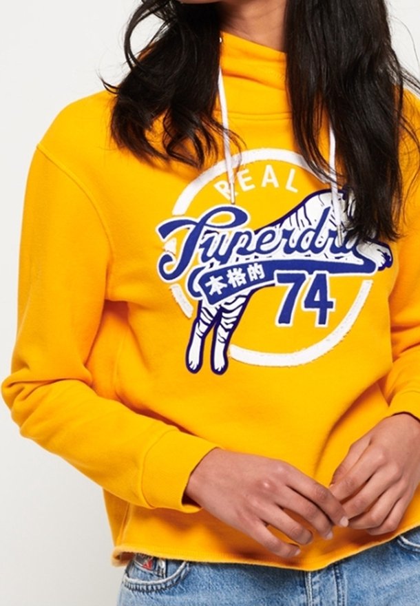 Personne portant un sweat à capuche jaune vif avec cordons blancs, logo "Superdry 74" bleu et blanc et graphisme tigre, associé à un jean bleu.