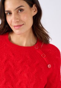 Donna con capelli castani che indossa un maglione rosso a trecce con tre bottoni rossi e dorati vicino al collo, che sorride dolcemente.