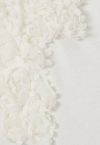 Weißer Stoff mit geschichteten, strukturierten floralen Applikationen an einer Kante, die sanfte, durchsichtige Blütenblätter und einen glatten Hintergrund aufweisen.