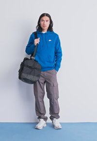 Fiatal ember kék Nike kapucnis pulóverben, szürke cargo nadrágban és fehér sportcipőben áll, fekete válltáskával egy sima háttér előtt.
