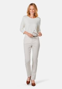 Lichte crème lange mouw top, aangesloten lichtbeige broek en bruine puntige hakken. Het model houdt een zonnebril vast en heeft middellang golvend haar.