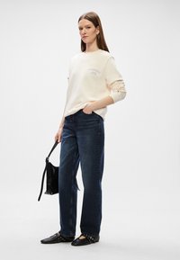 Ανοιχτό μπεζ φούτερ με μωβ γράμματα, συνδυασμένο με σκούρο μπλε τζιν σε ίσια γραμμή. Μαύρη τσάντα με λουράκι και επίπεδα μαύρα παπούτσια με τρουκ. 