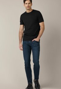 Czarna koszulka z krótkim rękawem, ciemnoniebieskie dopasowane dżinsy i czarne buty. Model stoi w pozycji wyprostowanej z rękami w kieszeniach na neutralnym tle.