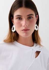 Store, skinnende, transparente hooop øreringe lavet af harpiks, der komplementerer en hvid top med en knude detalje nær halsudskæringen.