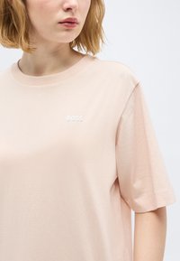 Νεαρή γυναίκα με κοντά ξανθά μαλλιά που φοράει ένα ανοιχτό ροζ φαρδύ μπλουζάκι με το λογότυπο "BOSS" στο στήθος σε απλό φόντο.