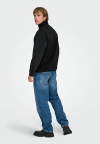 Giovane uomo in piedi di lato che guarda indietro, indossa una giacca nera, jeans blu e stivali neri su uno sfondo chiaro uniforme.