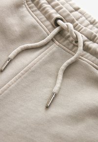 Pantaloni jogger beige con vita regolabile tramite lacci e punte in metallo, tessuto testurizzato e tasche laterali. Finitura liscia con dettagli di cucitura.