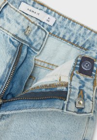 Jean en denim bleu clair avec une fermeture éclair, dotée d'un tirette en argent, de coutures brunes et d'une étiquette blanche à l'intérieur.