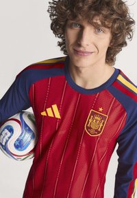 Crvena i plava Adidas majica sadrži grb, vertikalne pruge i žute akcentne detalje. Igrač drži šareni fudbalski lopti s uzorkom spirale.