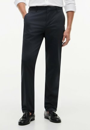 Mężczyzna w czarnych spodniach wizytowych z bocznymi kieszeniami i czarnych mokasynach z frędzlami, ręce swobodnie przy bokach, częściowo widoczna biała koszula.