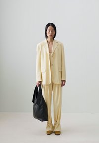 Yellow tailored suit with wide-legged trousers and a single-button blazer, paired with a black tote bag and yellow shoes. Smooth texture.