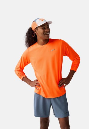Man die glimlacht met zijn handen op zijn heupen, gekleed in een feloranje longsleeve shirt, grijze shorts en een wit-oranje hardloopmuts voor een witte achtergrond.