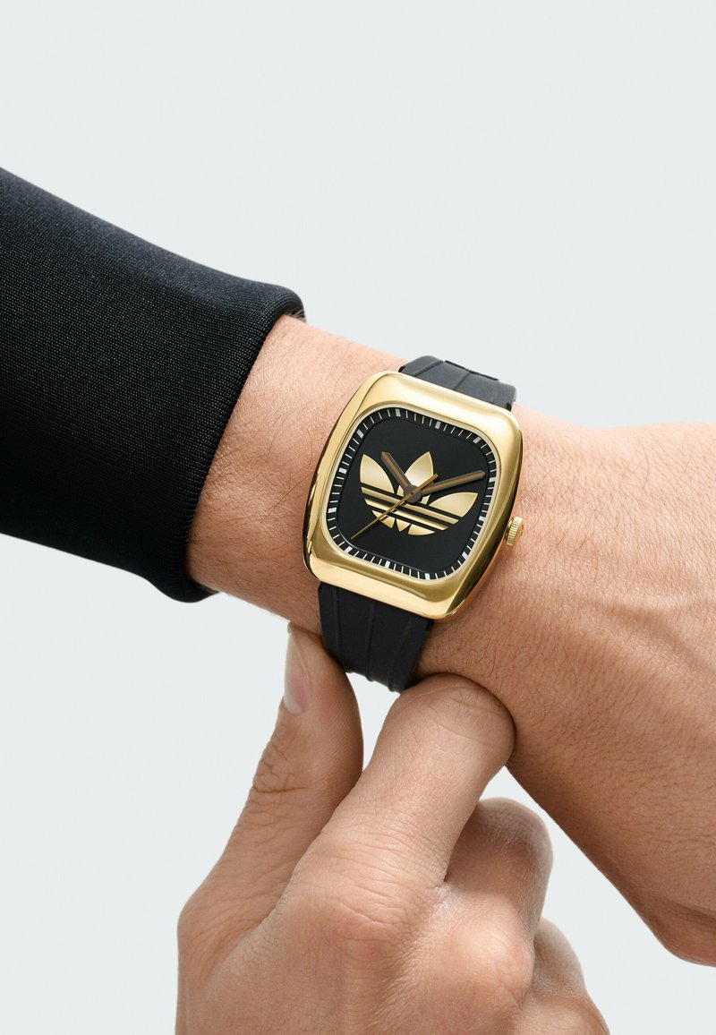 Handgelenk mit goldener und schwarzer quadratischer Uhr mit Adidas-Logo, das Armband mit der anderen Hand gegen einen hellen Hintergrund verstellend.