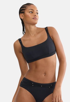 Woman with braided hair wearing a black asymmetrical bikini top and matching bikini bottom with small metallic accents.