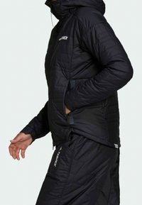 Sort isoleret jakke med quiltet mønster, med hætte, lynlåslommer og logo på brystet og venstre ærme.