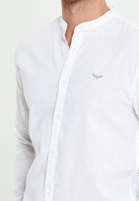 Chemise en lin blanc avec col mandarin, fermeture boutonnée à l'avant et un petit détail brodé gris sur la poitrine. Finition du tissu texturée.