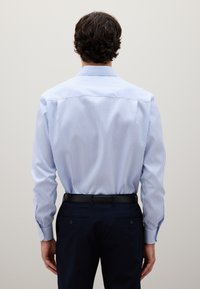 Hombre con cabello rizado oscuro vistiendo una camisa de vestir a rayas azul y blanca metida en pantalones negros, frente a un fondo liso.