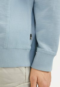 Hellblaues Sweatshirt mit langen Ärmeln, gerippten Bündchen und Saum, aus strukturiertem Stoff und mit einem kleinen Markenschild an der Seitennaht.