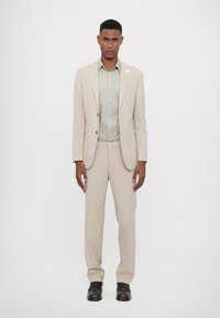 Completo beige a due pezzi con una vestibilità su misura, caratterizzato da una camicia verde chiaro sotto. L'insieme è completato con scarpe marroni scure.