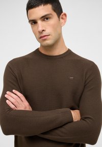 Pull en tricot marron avec une texture côtelée, col rond et un logo discret sur la poitrine. Il présente des manches longues et une coupe ajustée.