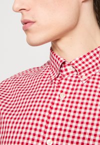 Chemise à carreaux rouge et blanc avec un col boutonné, présentant une texture lisse et de légers boutons sur la patte de boutonnage avant.