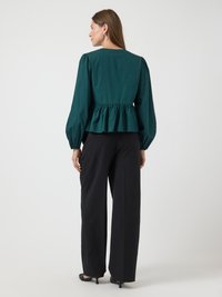 Blusa peplum verde con maniche lunghe a sbuffo, abbinata a pantaloni neri a vita alta e a gamba larga. Texture liscia, taglio semplice, dettagli minimi.