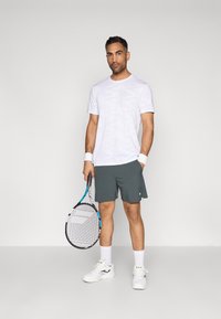 Λευκή μπλούζα με καμουφλάζ, γκρίζα σορτς, λευκά αθλητικά παπούτσια. Κρατάει μια ρακέτα τένις με μπλε λεπτομέρειες. Απλό φόντο.