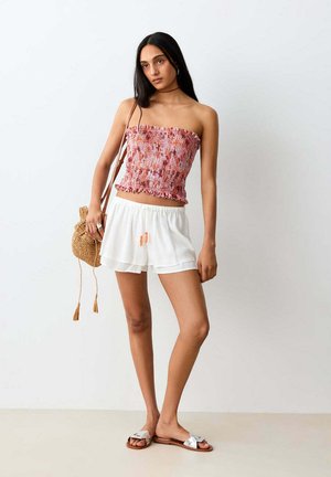 Femme aux longs cheveux foncés porte un top rose sans bretelles, un short blanc, des sandales argentées, et porte un petit sac tissé sur un fond uni.