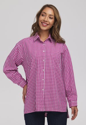 Kobieta uśmiechnięta, ubrana w luźną różowo-białą koszulę w kratę z długimi rękawami i guzikami, jedną ręką na biodrze, na jednolitym jasnym tle.