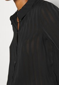 Blouse noire à rayures avec un devant à boutons, un petit col et une texture légère et transparente. Légère brillance du tissu.