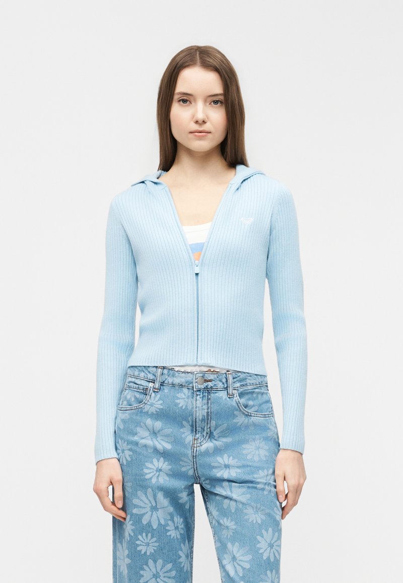 Νεαρή γυναίκα που φοράει ένα ανοιχτό μπλε ριμπ ζιπ-απ φούτερ και ψηλόμεσα μπλε τζιν με μεγάλο λουλουδάτο μοτίβο, στέκεται μπροστά σε μια απλή φόντο.