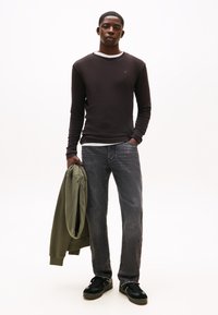 Camicia marrone scuro a maniche lunghe sopra una maglietta bianca a girocollo, jeans grigi con trama, scarpe da ginnastica nere e giacca verde oliva tenuta in mano.