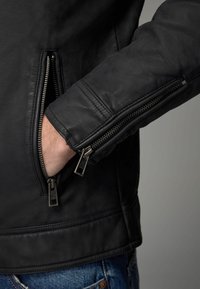 La chaqueta de cuero negro cuenta con bolsillos y puños con cremalleras, tiene una textura suave y una silueta estructurada, con costuras detalladas en el dobladillo.