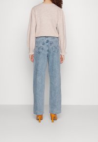 Femme portant un jean bleu clair droit, un haut beige à manches longues et des chaussures à talons hauts orange, debout de dos sur un fond uni.