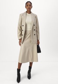 Beige Kunstleder-Blazer mit schwarzen Knöpfen, kombiniert mit einem passenden Midirock. Das Model trägt schwarze Stiefeletten und hält eine kleine schwarze Tasche.