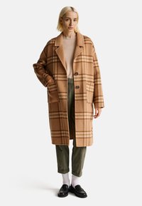 Beżowy wełniany płaszcz w kratkę o luźnym kroju, z klapami, dwoma guzikami i bocznymi kieszeniami. Połączony z zielonymi spodniami z korduroju i czarnymi mokasynami.